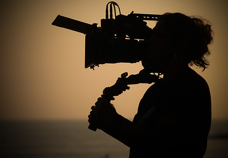 Silhouette of a camerawoman with a camera mounted on her shoulder, with the sea and sky blurry in the background